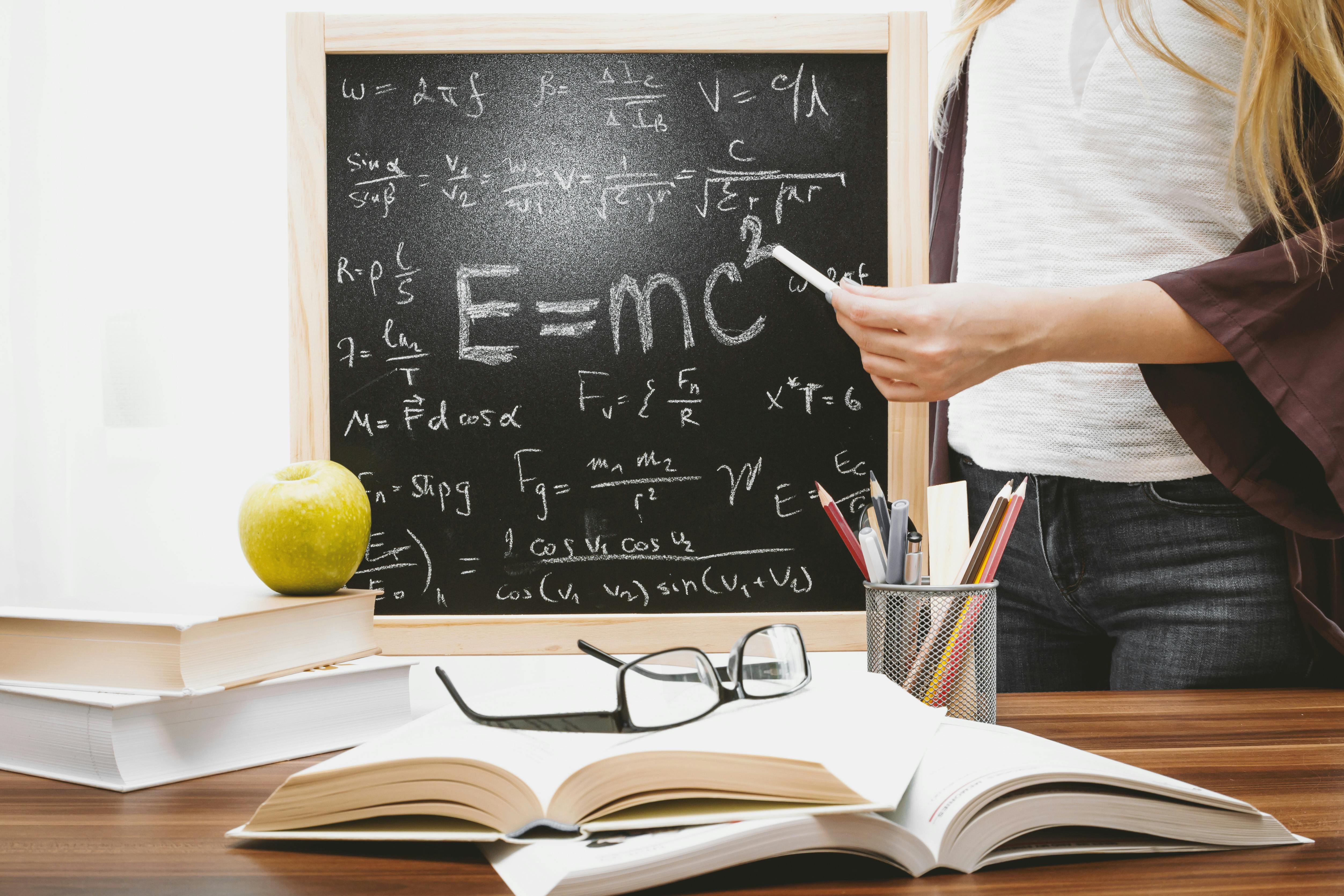 Person Standing With Books On A Table And A Blackboard In The Background